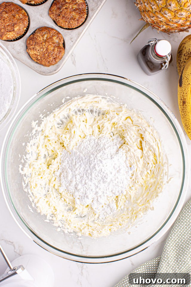 Overhead view of powdered sugar being gradually added to a bowl of creamy beaten butter and cream cheese for frosting.