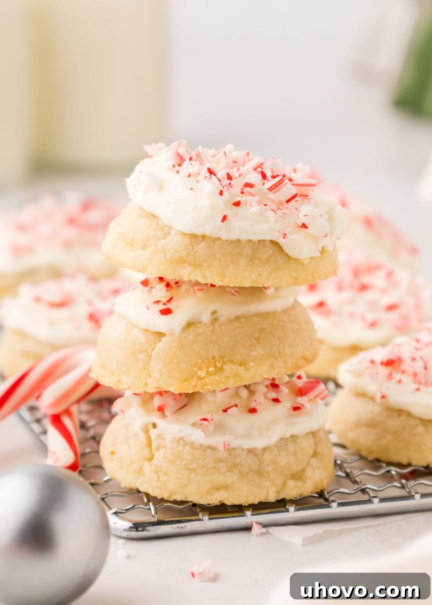 A stack of three frosted peppermint meltaways, adorned with crushed candy canes, with more cookies softly blurred in the background, on a wire rack.