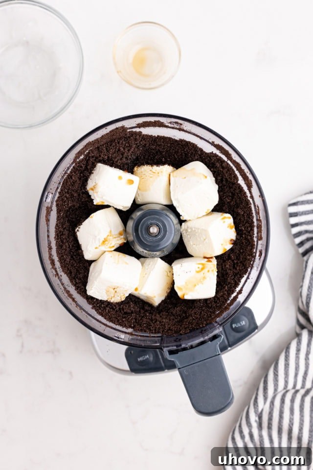 Overhead view of cream cheese and vanilla extract being added to finely crushed Oreo crumbs in the bowl of a food processor.
