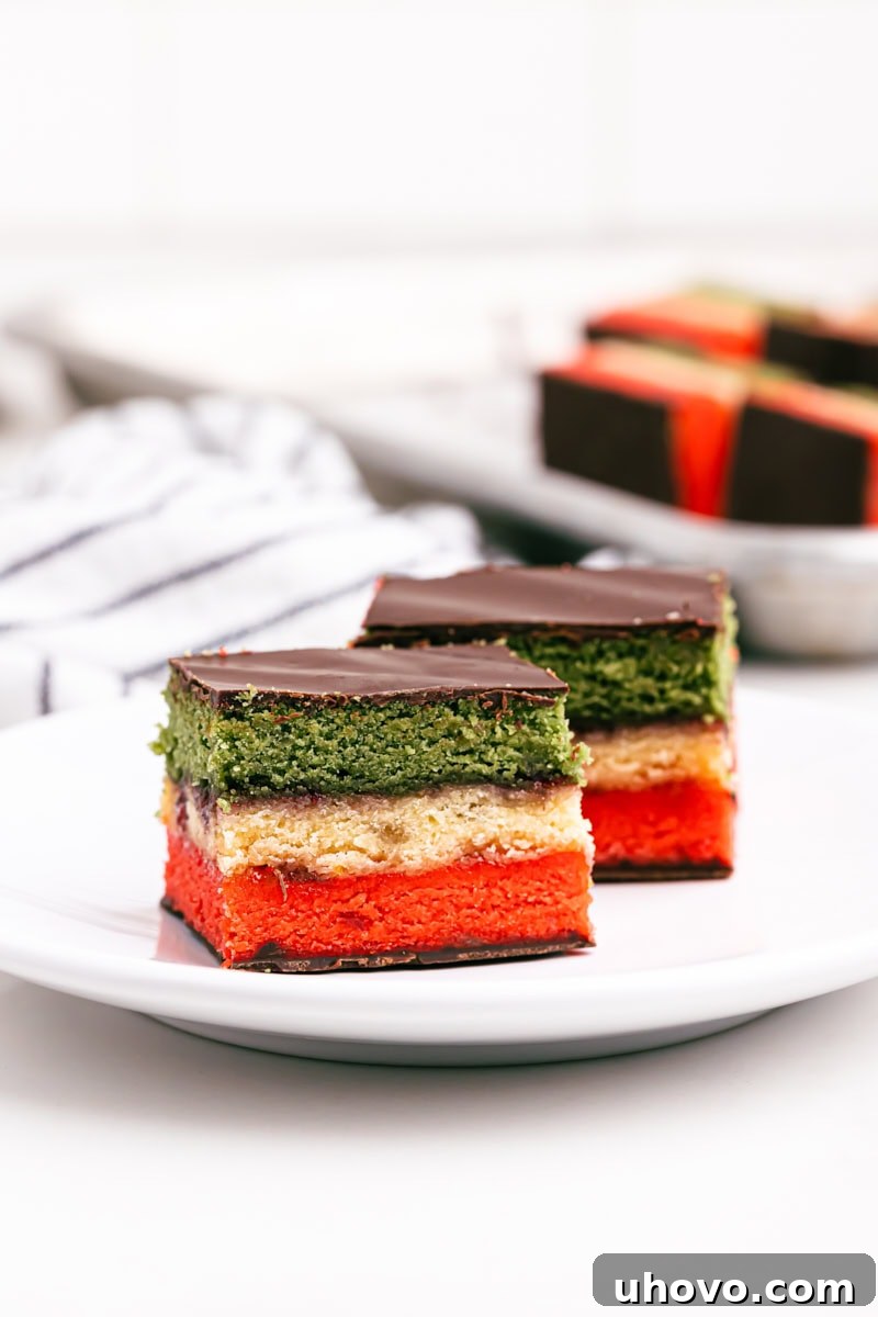 Two Italian rainbow cookies on a white plate with more cookies in the background, showcasing their vibrant layers.