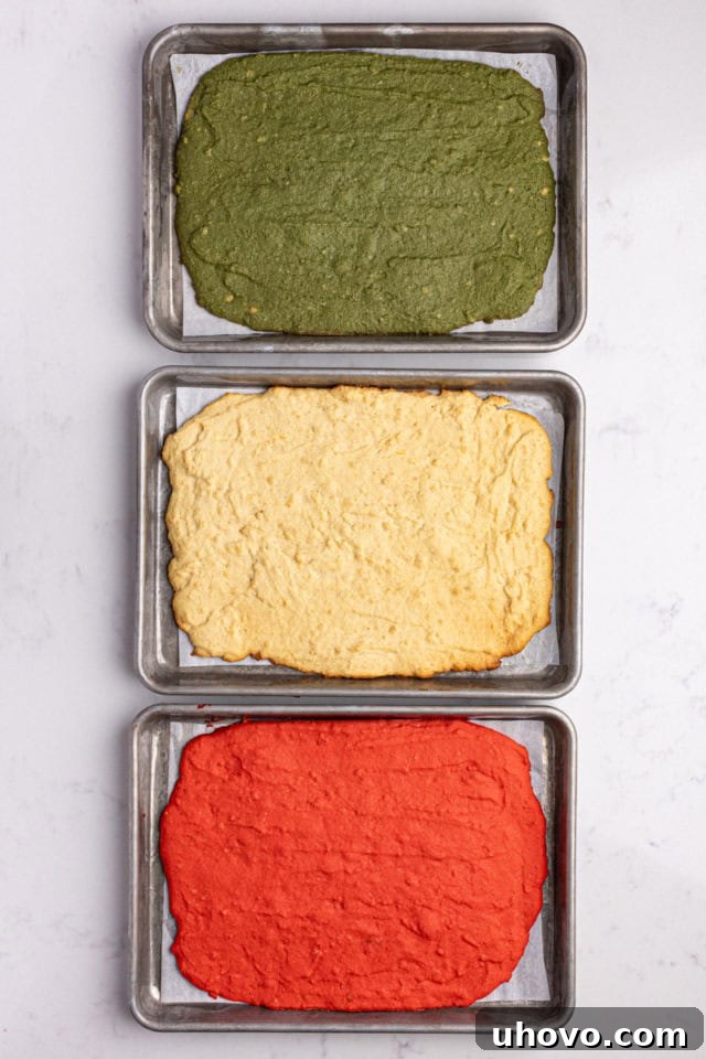 Overhead view of three baked portions of Italian rainbow cookie dough in different colors (red, green, and plain) on baking sheets.