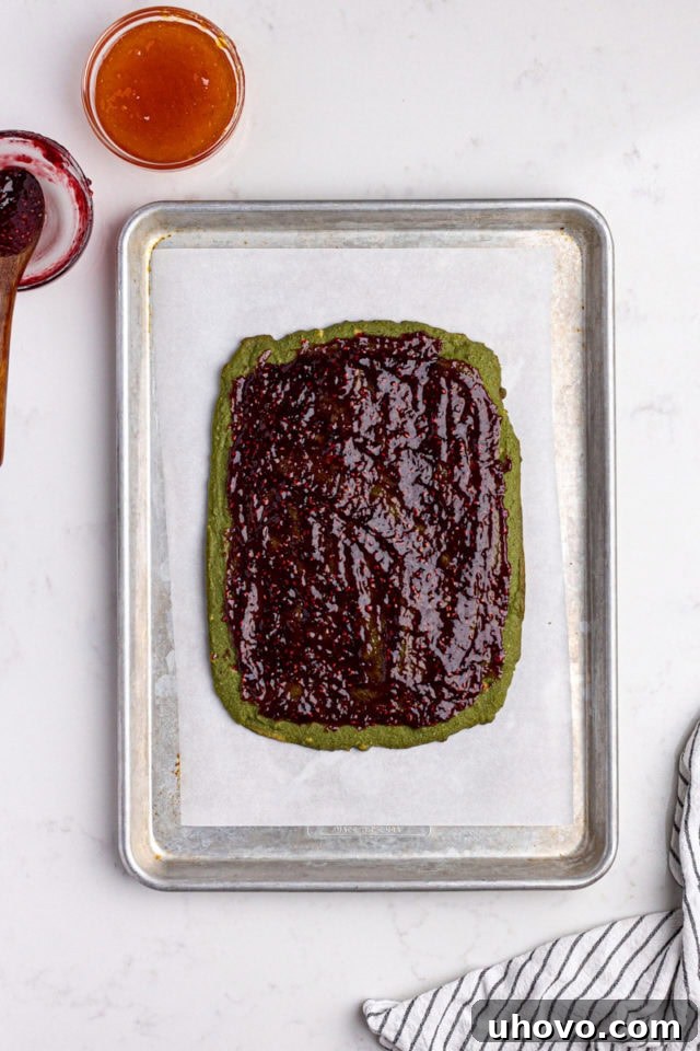 Overhead view of raspberry preserves being spread thinly and evenly over the green layer of Italian rainbow cookies.