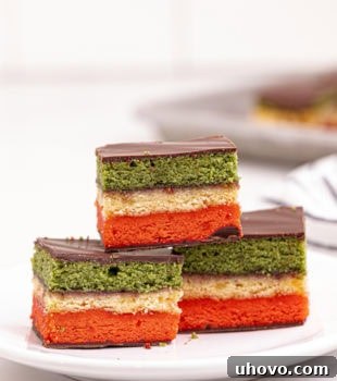 An Italian rainbow cookie on top of two more cookies on a white plate, showing its layers and chocolate.