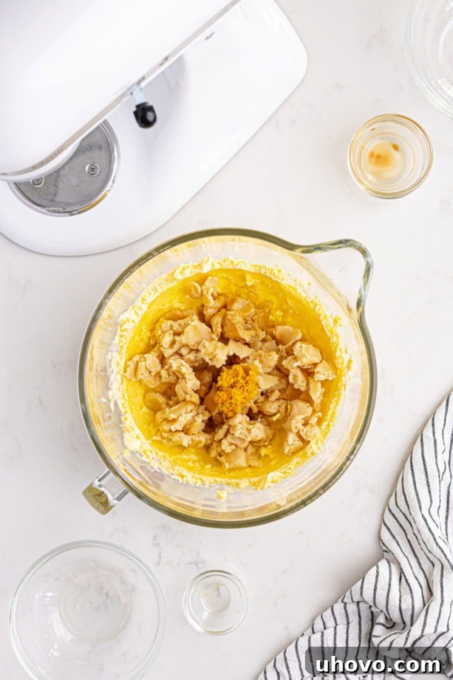 Overhead view of almond paste, almond extract, vanilla extract, and orange zest being added to the creamed butter and sugar mixture.