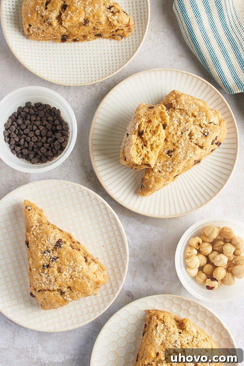 Chocolate chip-hazelnut scones arranged on white patterned plates, with small bowls of chocolate chips and hazelnuts in the background.