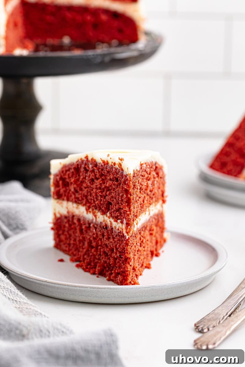 A beautifully presented slice of red velvet cake on a white plate, with the remainder of the cake in the background, showing the layered perfection.