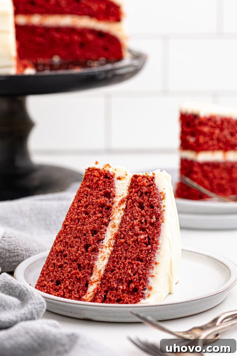 A delicious slice of red velvet cake on a white plate, with the rest of the frosted cake blurred in the background, inviting you to enjoy.