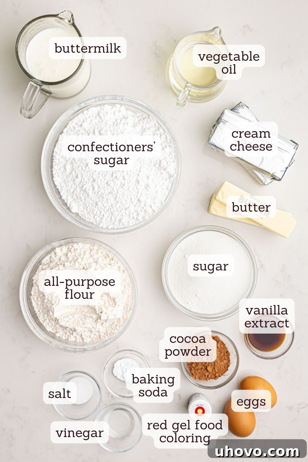 An overhead view of all the essential ingredients laid out for baking a red velvet cake, including flour, sugar, cocoa, buttermilk, oil, eggs, and food coloring.