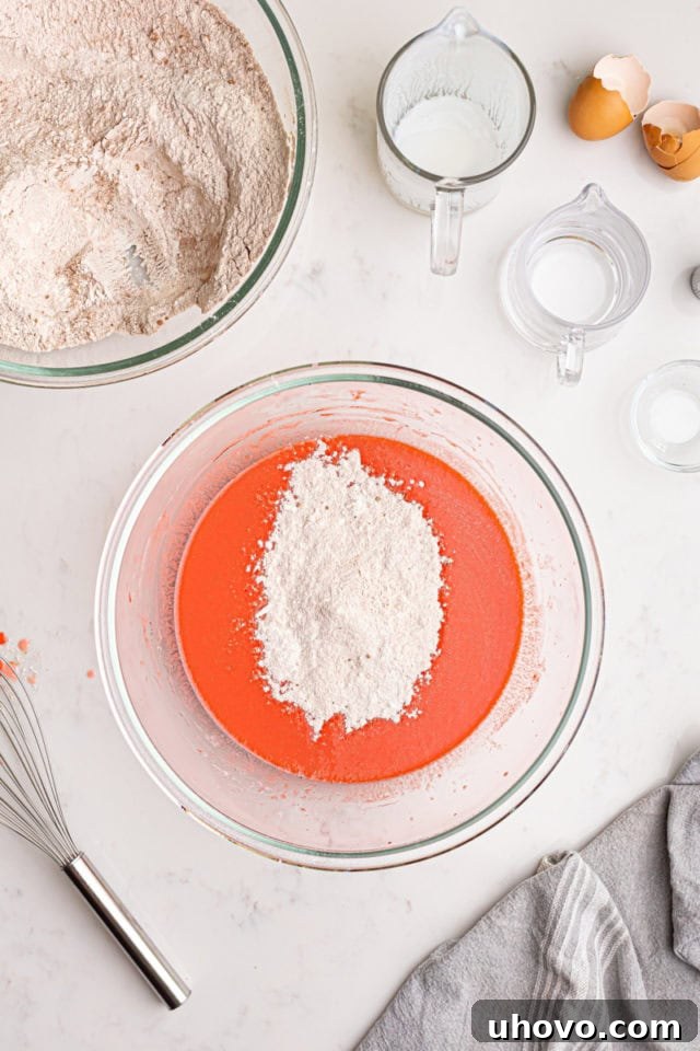 The dry ingredients for red velvet cake being gradually added to the wet ingredients in a mixing bowl, showing the beginning of the batter formation.