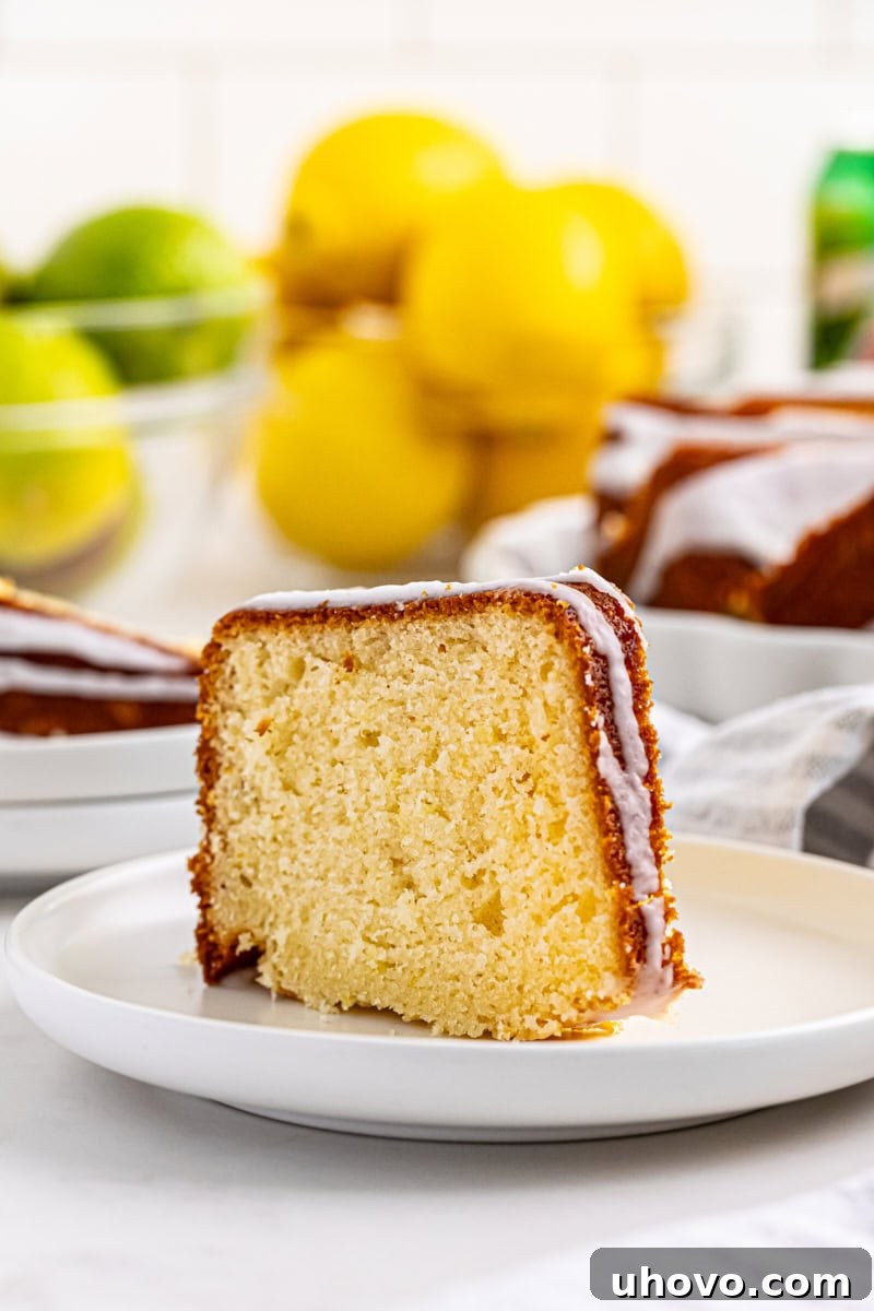 A perfectly portioned slice of 7UP cake on a white plate, garnished with a lemon slice, with limes and lemons blurred in the background.