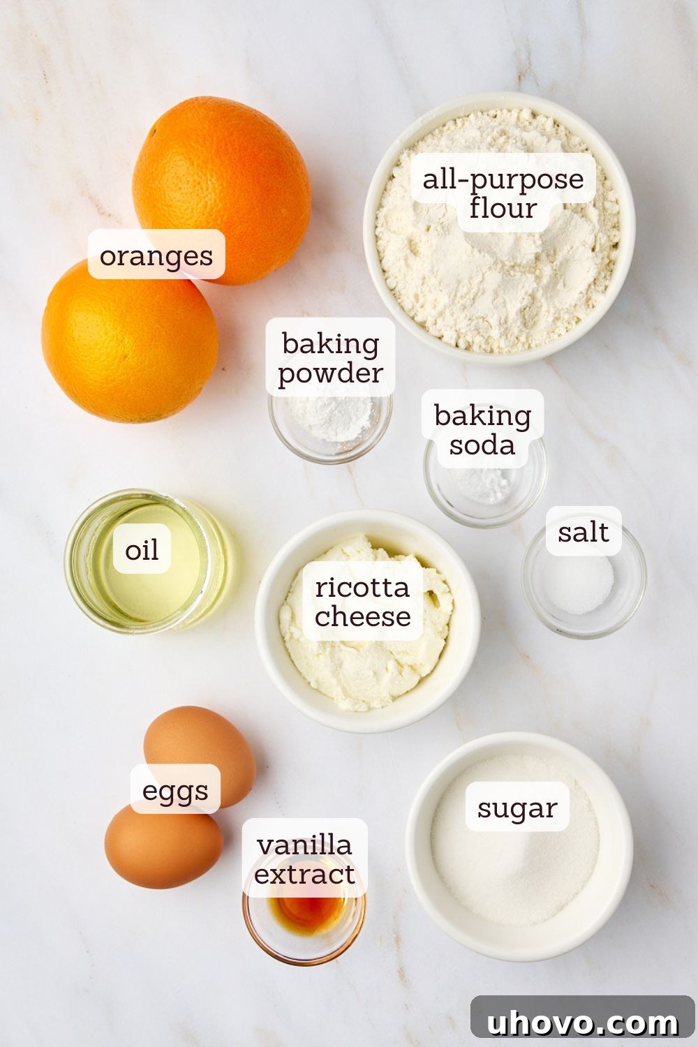 Overhead view of various fresh ingredients laid out on a clean surface, including flour, sugar, fresh oranges, eggs, and a bowl of ricotta cheese, ready for making orange ricotta muffins