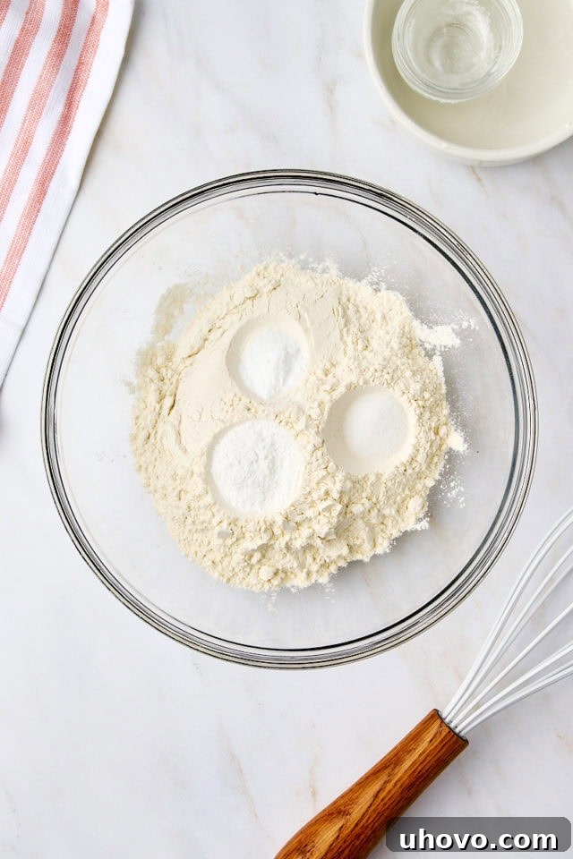 Overhead view of flour, baking powder, baking soda, and salt measured into a clear glass mixing bowl, awaiting whisking