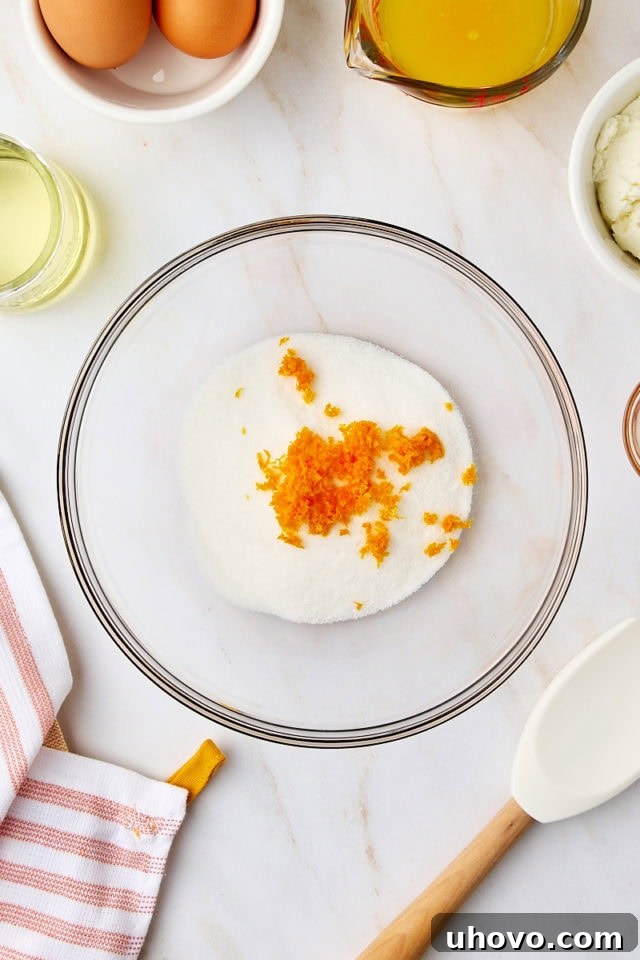 Overhead view of granulated sugar and vibrant orange zest in a glass mixing bowl, ready to be rubbed together