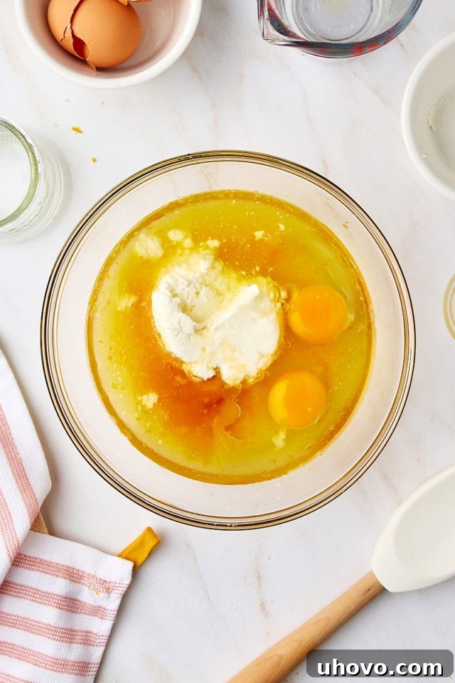 Overhead view of orange juice, ricotta cheese, vegetable oil, eggs, and vanilla extract being added to the sugar and zest mixture in a glass mixing bowl