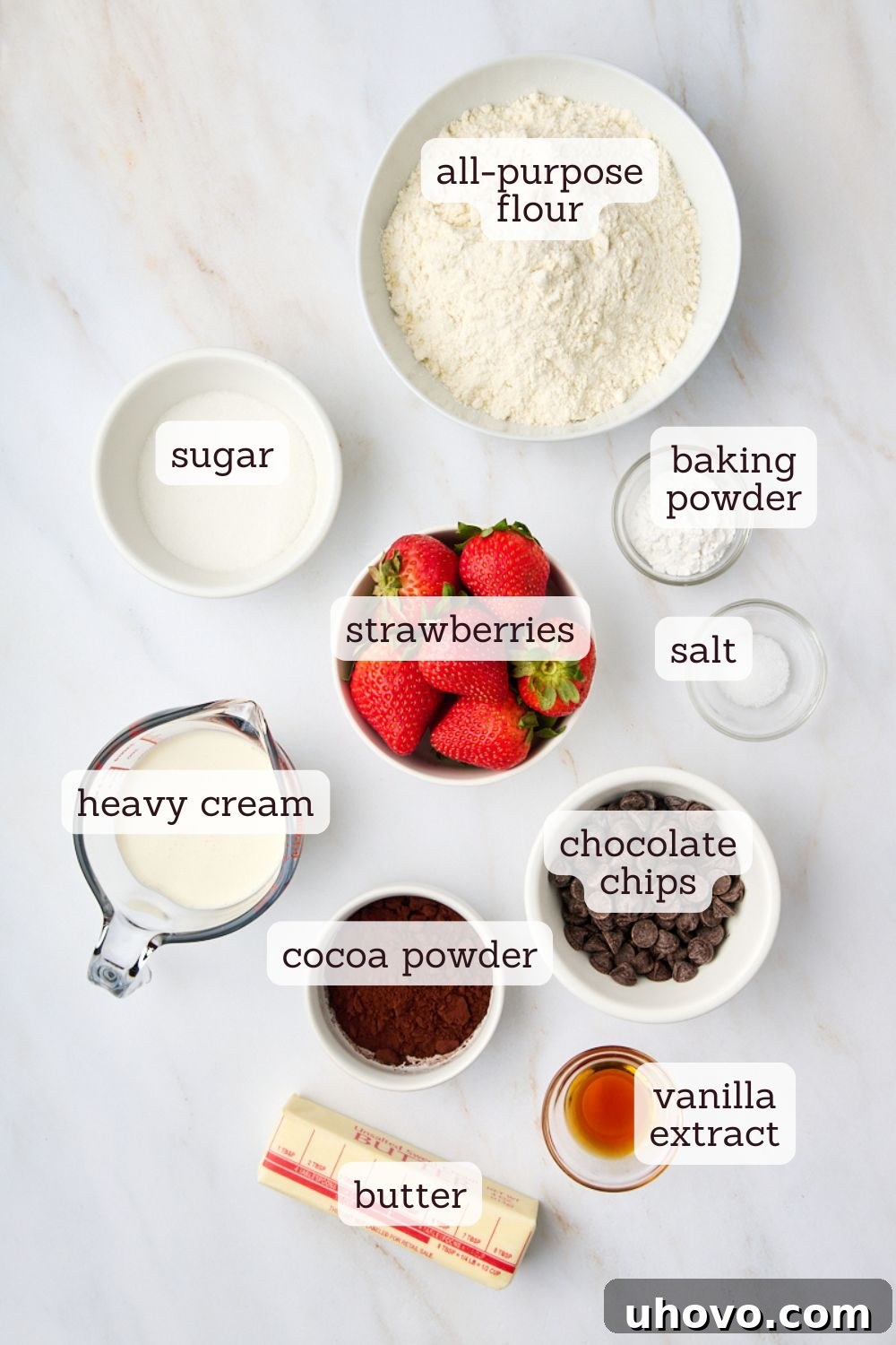 Overhead view of all the raw ingredients for chocolate strawberry scones laid out on a white surface, including flour, sugar, cocoa powder, butter, strawberries, chocolate chips, cream, and vanilla extract.