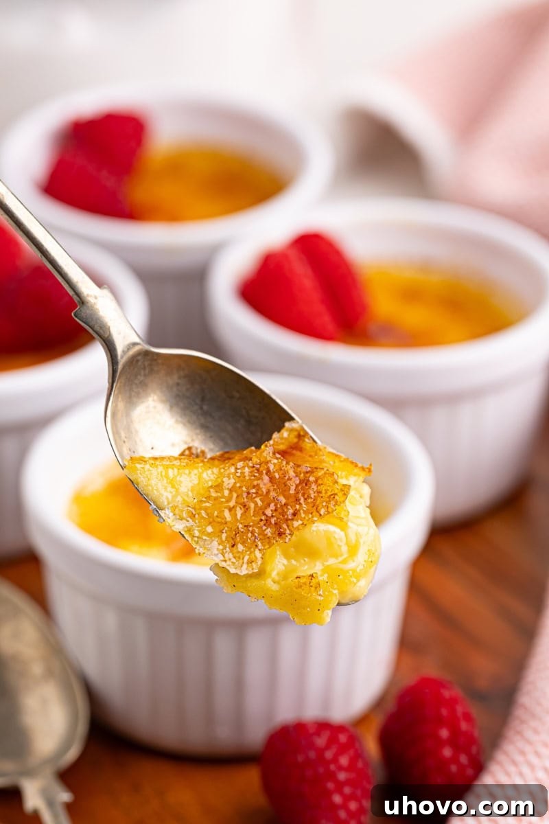 A close-up shot of a spoon breaking through the crisp caramelized sugar topping of a crème brûlée, with more ramekins blurred in the background, highlighting the perfect texture.