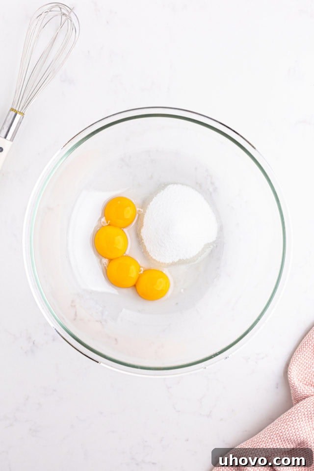 An overhead view of golden egg yolks and granulated sugar being whisked together in a mixing bowl until light and well combined.