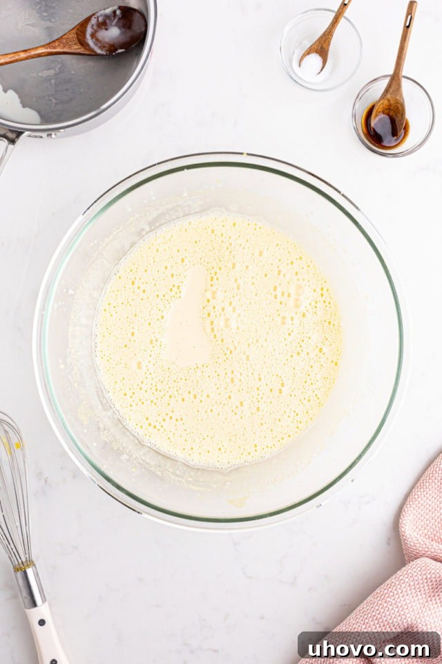 An overhead shot showing warm heavy cream being slowly and carefully poured into a bowl containing the whisked egg yolks and sugar, while continuously whisking to temper the eggs.