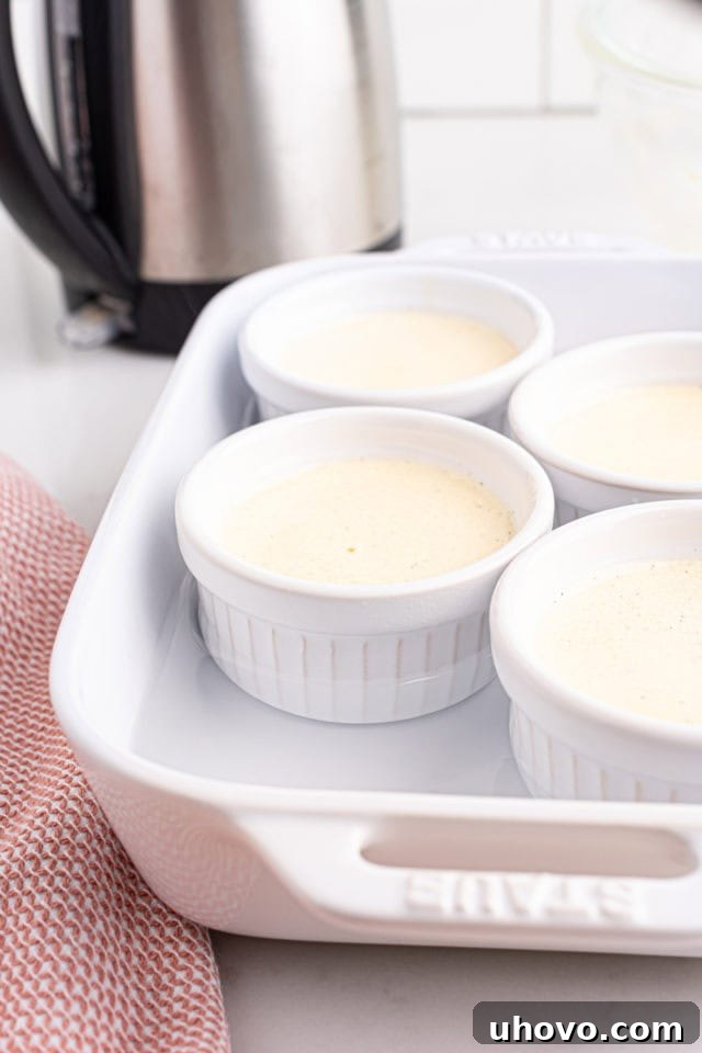 Four white ceramic ramekins, evenly filled with the smooth crème brûlée custard mixture, are placed inside a larger baking pan, with hot water carefully poured halfway up their sides for a water bath.