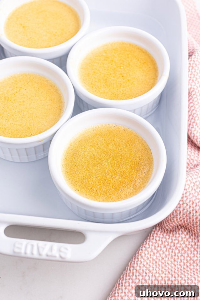 Four freshly baked crème brûlée ramekins, showing their perfectly set but slightly wobbly centers, are resting within the baking pan after being removed from the oven.
