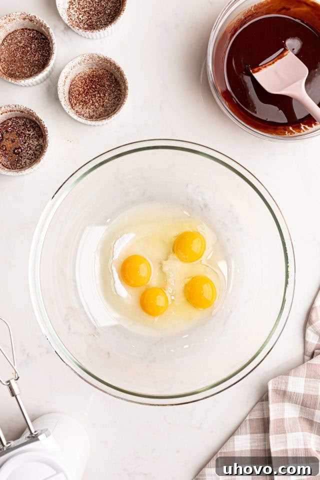 An overhead perspective of a glass mixing bowl containing eggs, granulated sugar, and a pinch of salt, being whisked vigorously to create a light and frothy mixture.