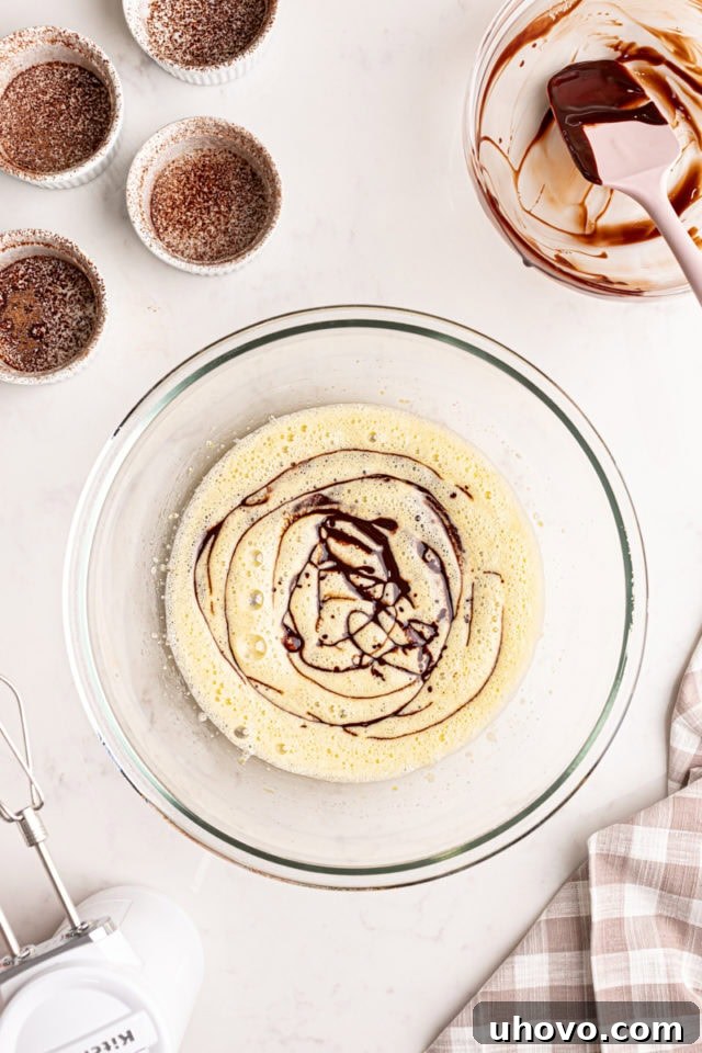 An overhead shot showing the cooled, melted chocolate and butter mixture being carefully poured into the light and frothy egg and sugar mixture in a glass bowl.