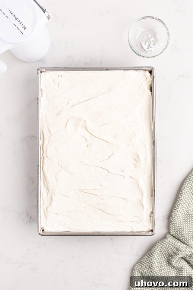 Overhead view of a generous layer of fluffy whipped cream topping being spread smoothly over the chilled tres leches cake in a baking pan.