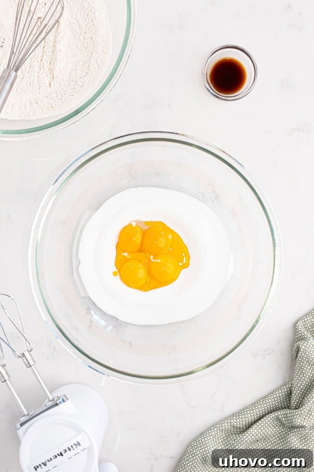 Overhead view of vibrant yellow egg yolks and granulated sugar actively being beaten together in a glass mixing bowl, showing a pale and fluffy texture.