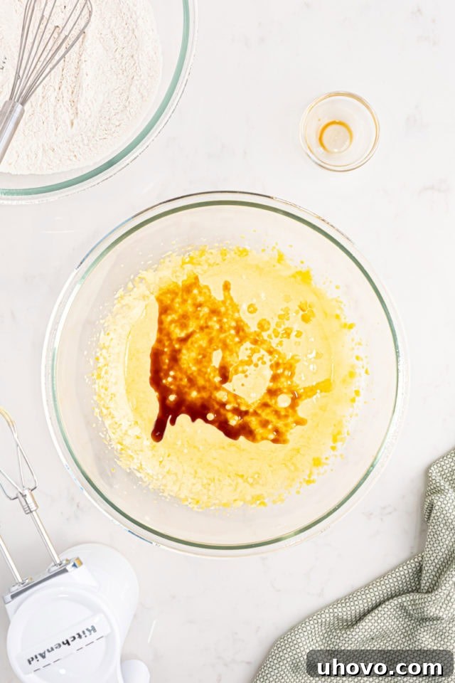 Overhead view of whole milk and vanilla extract being poured into the pale, fluffy egg yolk and sugar mixture in a mixing bowl.
