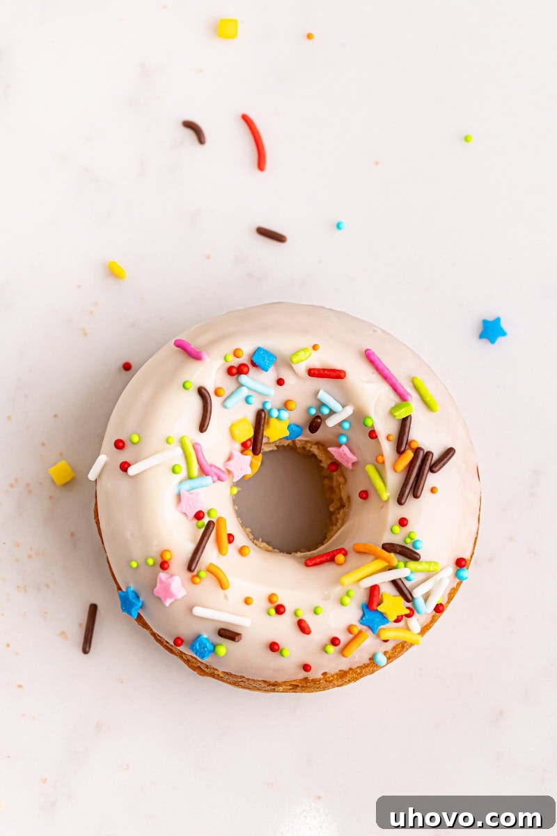A single baked doughnut with white glaze and colorful sprinkles sits invitingly on a white surface.
