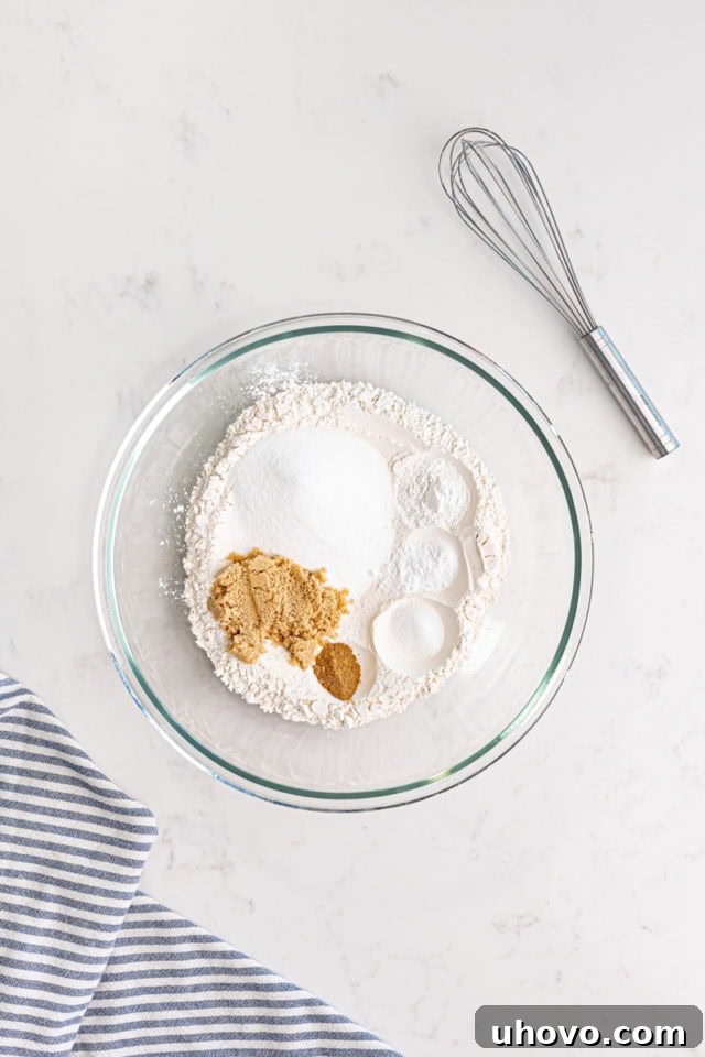 Overhead view of flour, sugar, brown sugar, baking powder, baking soda, salt, and nutmeg in a glass mixing bowl, representing the dry ingredients.