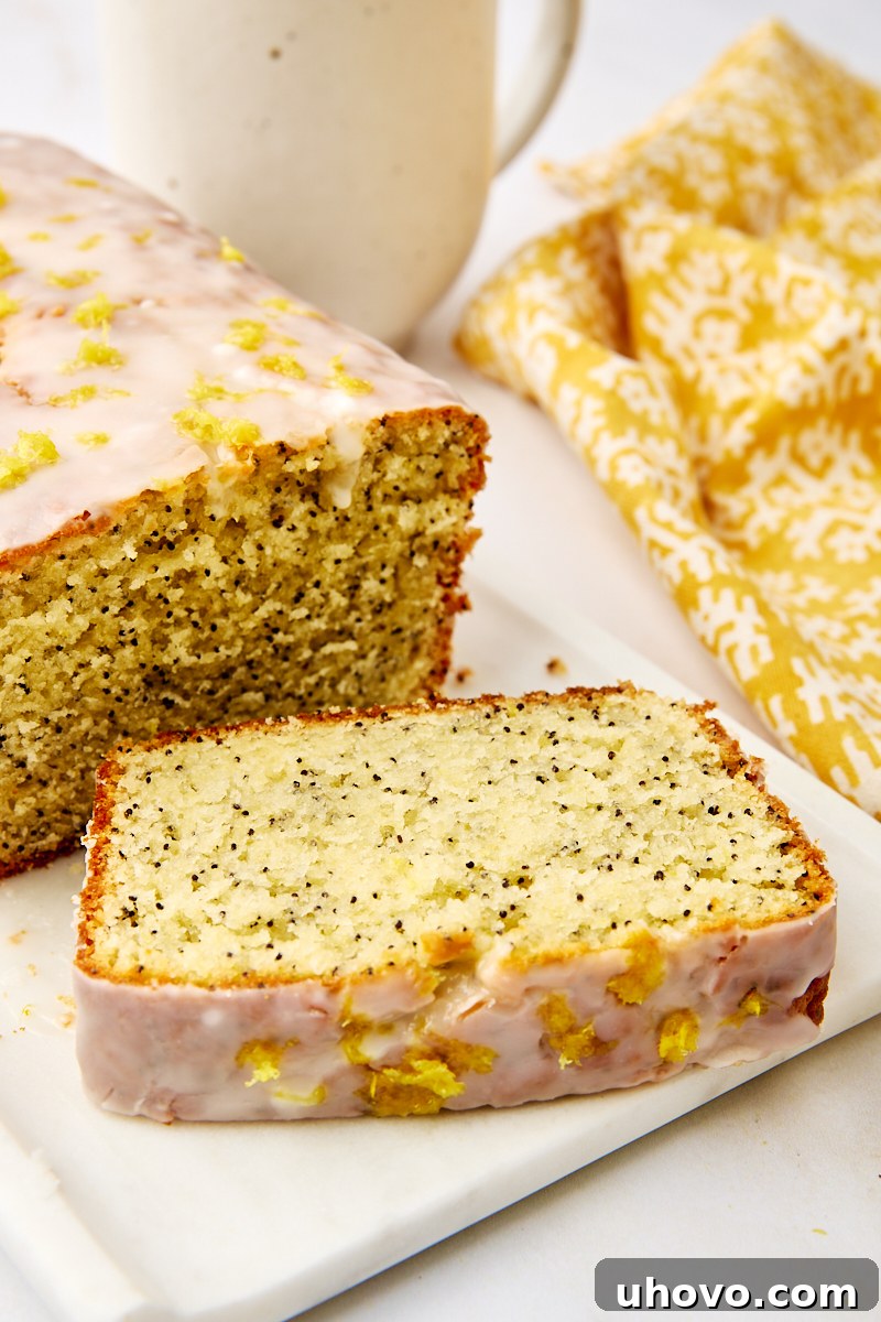 A perfectly sliced lemon poppy seed loaf on a plate, showing its moist texture and speckled poppy seeds, with the remaining loaf in the background.