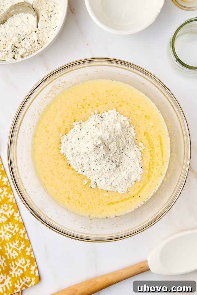 An overhead shot showing the gradual addition of the dry flour mixture into the wet lemon-infused ingredients, being gently folded to create the loaf batter.