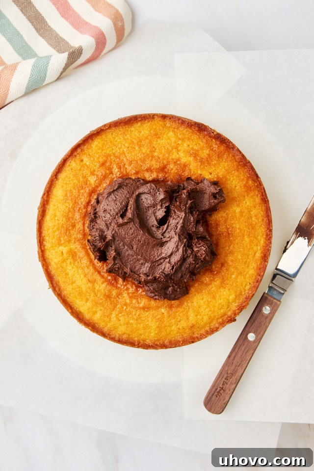 A generous layer of smooth chocolate frosting being spread evenly on top of a cooled yellow cake layer, preparing for the second layer.