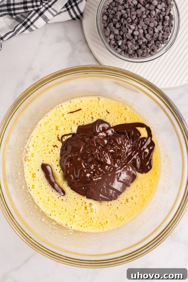 An overhead view of the rich, melted chocolate mixture being carefully poured into a bowl containing the whisked sugar and egg mixture, about to be combined with vanilla.