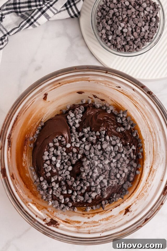 A vibrant overhead shot illustrating mini chocolate chips being folded into the deep chocolate cookie batter in a glass mixing bowl, adding delightful pockets of sweetness.