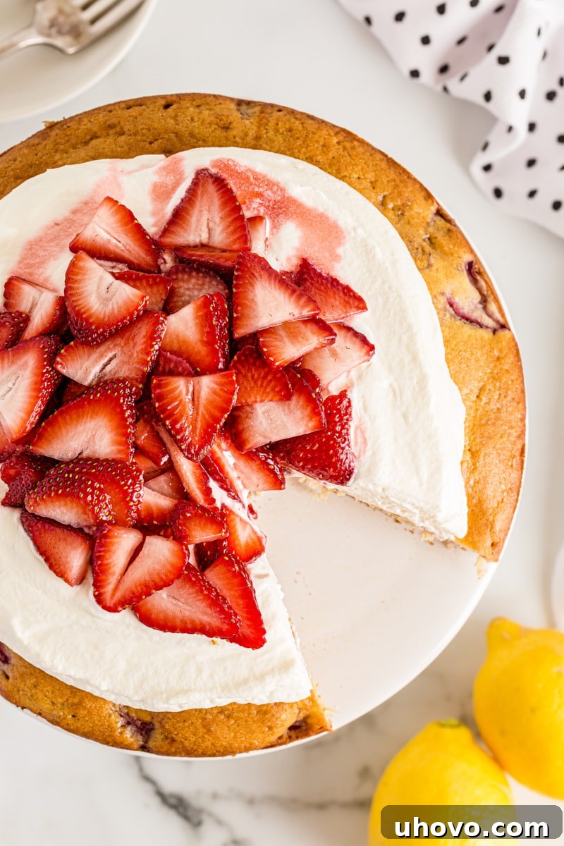 An overhead view of a sliced Lemon-Strawberry Shortcake, with one slice elegantly removed, revealing the moist cake, whipped cream, and fresh strawberry layers.