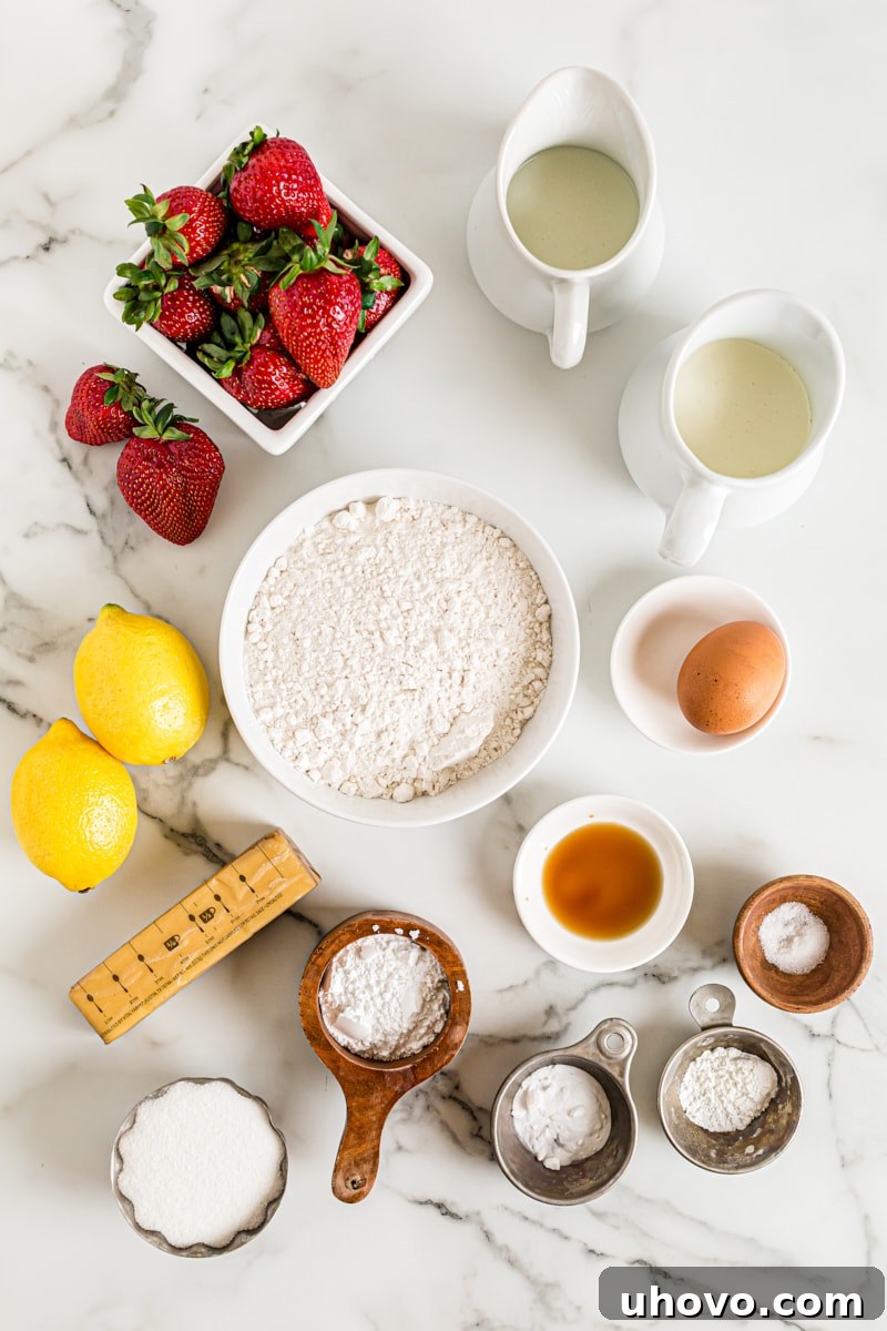An overhead view of all the fresh ingredients for Lemon-Strawberry Shortcake: flour, sugar, eggs, butter, lemons, and a bowl of vibrant strawberries.