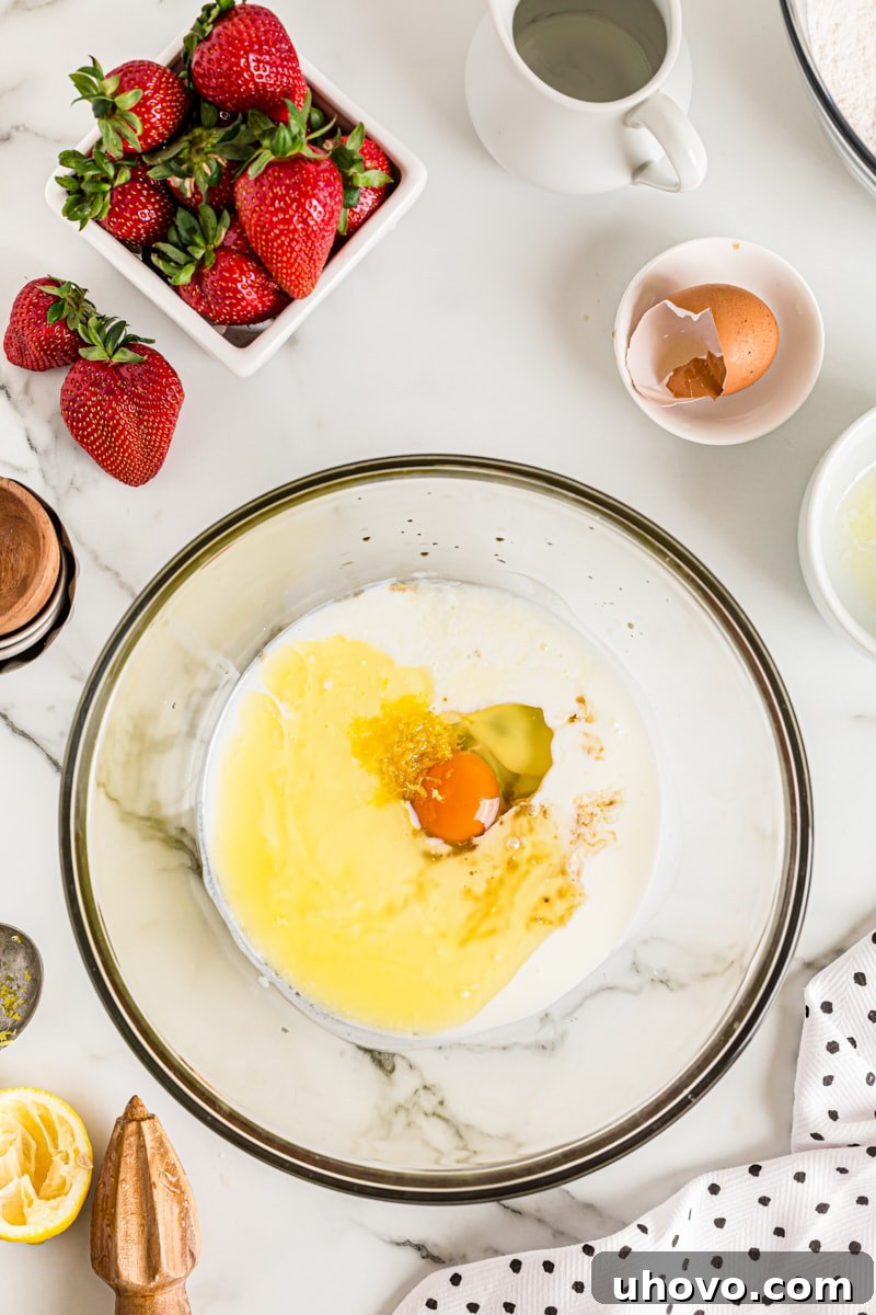 An overhead view of the wet ingredients for the cake (egg, buttermilk, melted butter, lemon juice, lemon zest, vanilla extract) in a separate glass mixing bowl, ready to be combined.