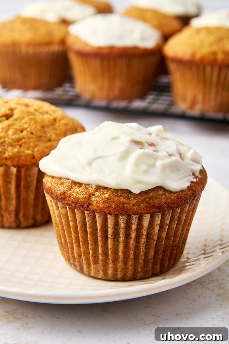 Close-up of a carrot cake muffin topped with a luscious cream cheese glaze on a white plate. Several unfrosted carrot cake muffins rest nearby on a cooling rack.