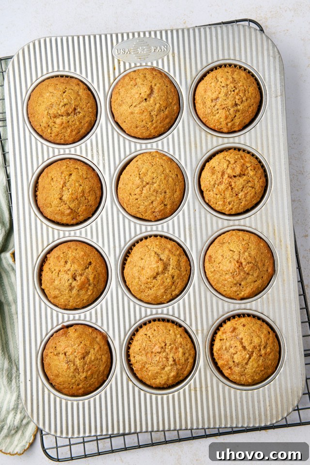 A metal muffin tin with twelve evenly baked carrot cake muffins sits on a cooling rack. Each muffin boasts a golden-brown top. A green cloth is partially visible on the side, set against a light countertop backdrop.