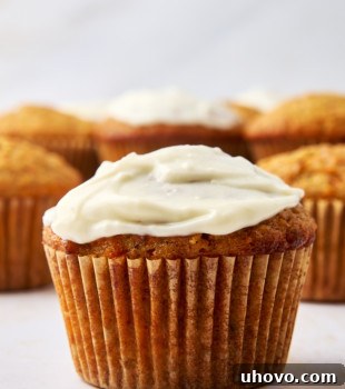 A close-up of a frosted cupcake with a luscious cream cheese glaze in the foreground, while a few unfrosted carrot cake muffins blur softly in the background. The frosting is creamy and white, contrasting with the golden brown cupcakes set on a light textured surface.