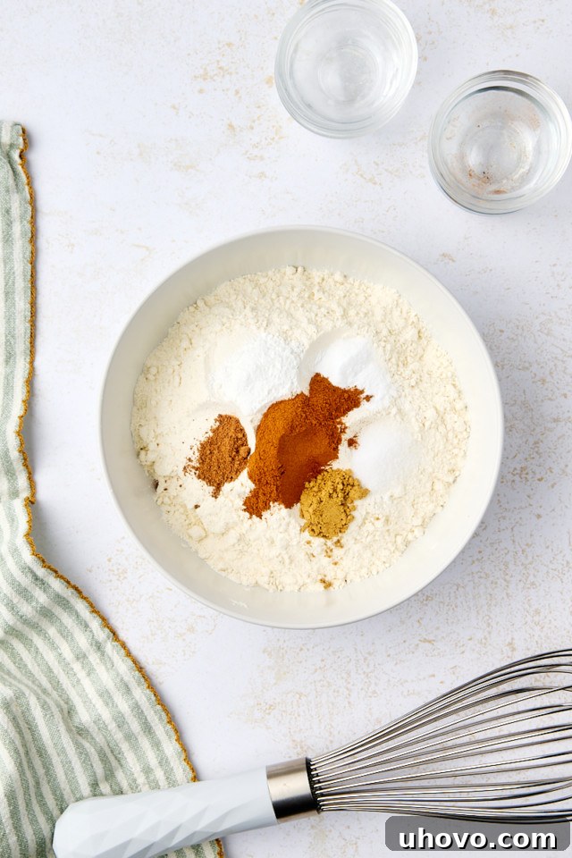 A white bowl containing flour, cinnamon, ginger, and baking powder on a light surface. A whisk with a white handle is nearby, alongside a green-striped cloth and two small glass bowls.