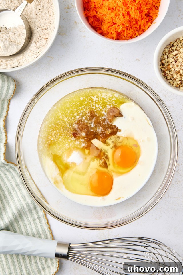 A glass bowl with eggs, milk, and other ingredients for a batter. Surrounding it are bowls of grated carrots, chopped nuts, flour, and a measuring cup. A whisk and a striped cloth are nearby on the white surface.