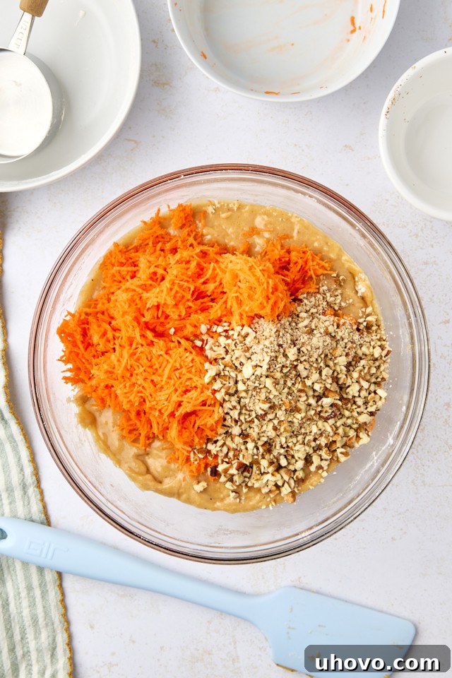 A glass bowl containing a mixture of shredded carrots, chopped nuts, and batter. Surrounding the bowl are several empty dishes, a measuring cup, and a blue spatula on a white surface. A striped cloth is partially visible on the left.