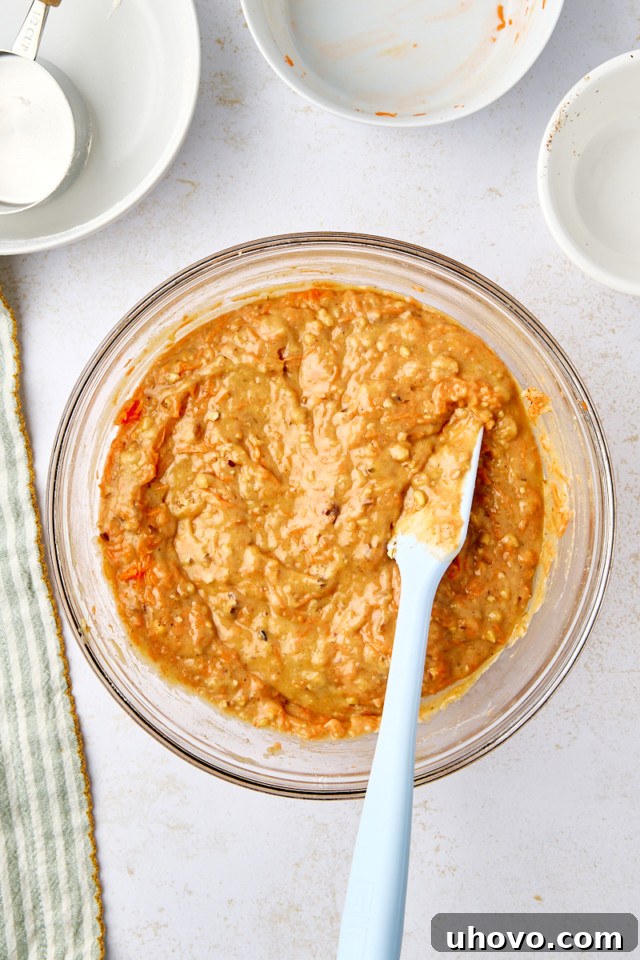 A glass bowl filled with thick, beige carrot cake muffin batter. A blue spatula rests inside the bowl. Nearby are empty white bowls, a measuring cup, and a striped cloth on a white surface.