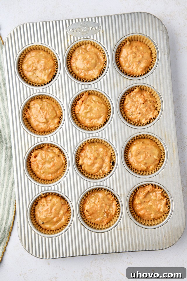 A muffin tray filled with 12 unbaked carrot cake muffins, each in a paper liner. The golden-brown muffins are evenly distributed and rest on a light-colored surface. A striped cloth is partially visible on the side, adding a touch of charm.
