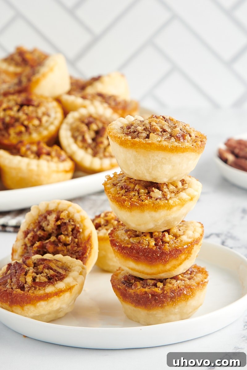 A stack of four mini pecan pies on a plate next to two other mini pies, showcasing their golden-brown crust and glistening pecan topping.