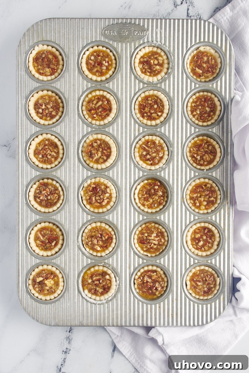 A mini muffin tin filled with unbaked pie crusts, each meticulously filled almost to the brim with the rich pecan pie mixture, ready for the oven.