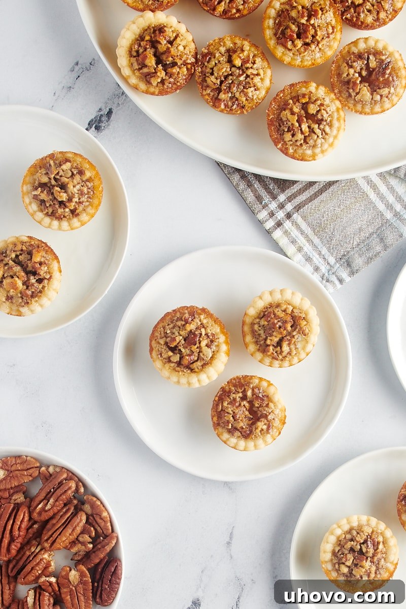 Three perfectly baked mini pecan pies elegantly arranged on a white plate, with a larger platter of pies blurred in the background, ready for serving.